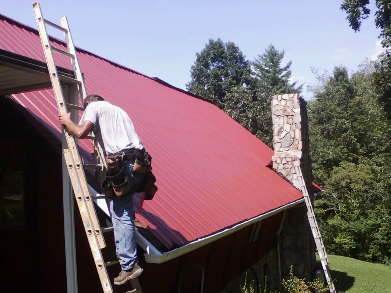 Roofer on a ladder beside a red metal roof and stone chimney — Soffit & Fascia Repair by Roofing Brothers in Tekoa
